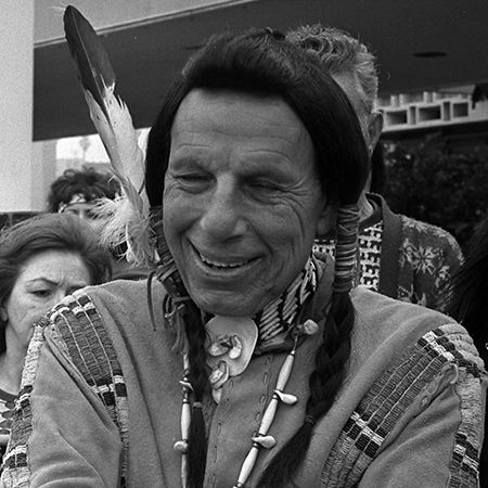 A middle-aged Iron Eyes Cody, dressed in a traditional Native American cloak and with a feather on his head, in an undated publicity photo