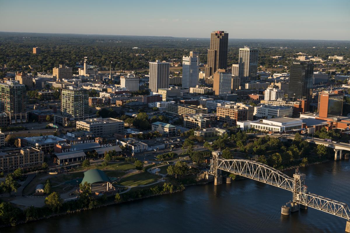 An aerial photo of Little Rock, Arkansas. A metal bridge over a river is in the foreground, skyscrapers are in the middle of the picture and forests in the background.